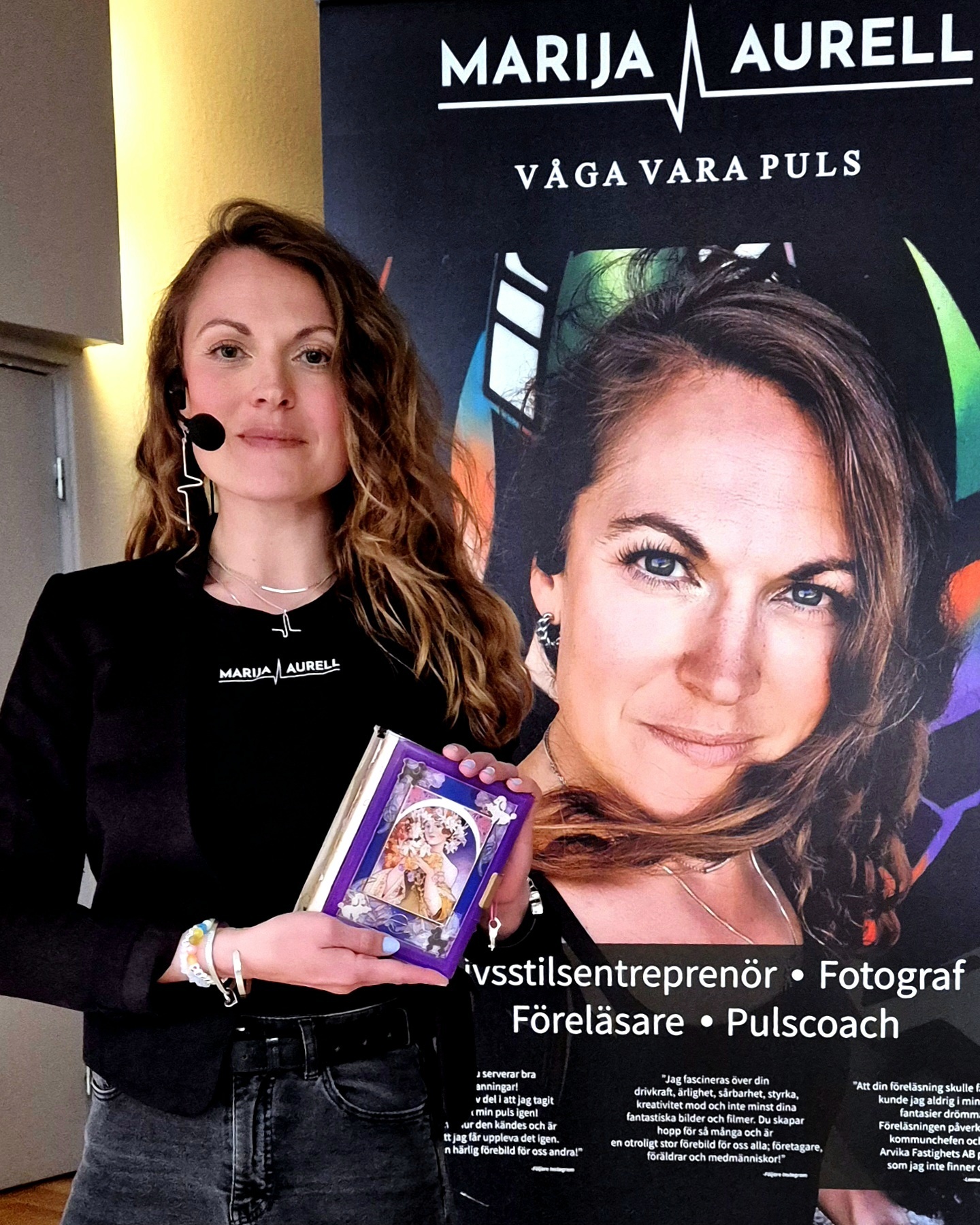 Marija står vid sin banner med Våga Vara Puls och håller sin bok